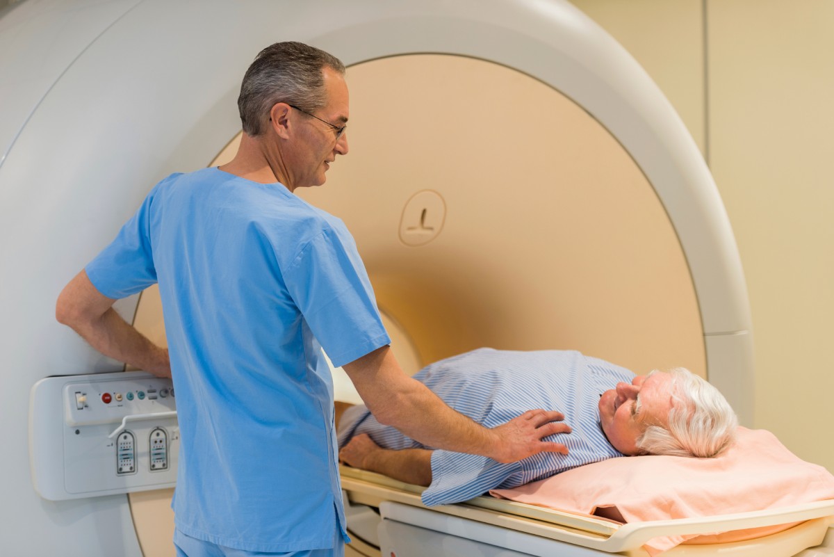 Un técnico está de pie al lado de un hombre acostado sobre una mesa que se desliza dentro de un dispositivo con forma de túnel.