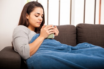 Young woman sitting on a sofa wrapped in a blanket and holding a large mug.