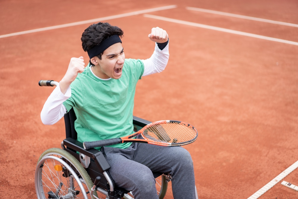 Un adolescente en una silla de ruedas con una raqueta de tenis en el regazo y con los brazos levantados celebra una victoria en la cancha de tenis.