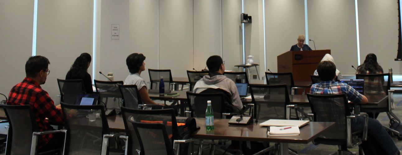 Fellows sitting at desks and listening to a speaker at a lectern.