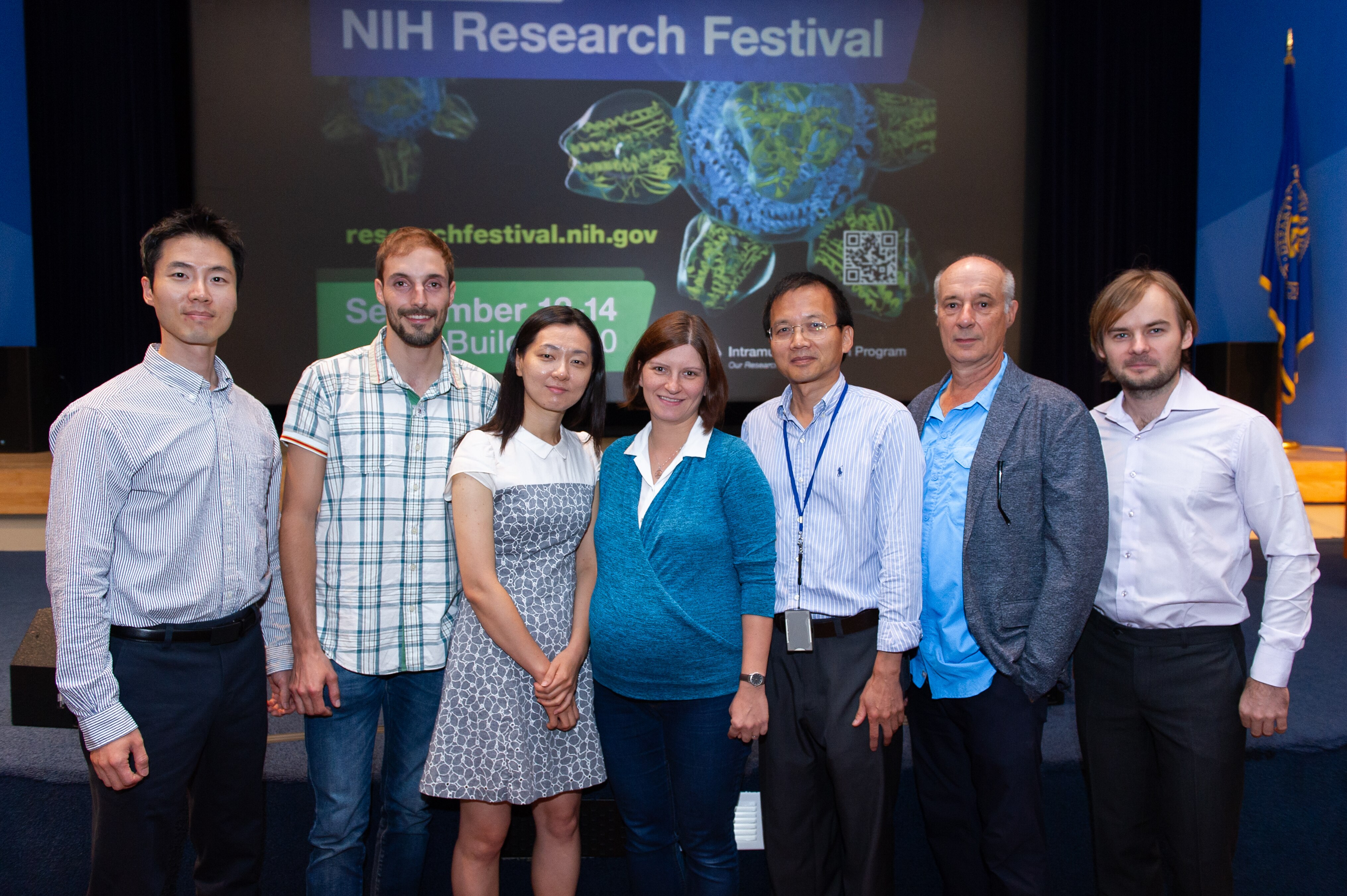Laboratory of Genetics and Physiology team members standing and smiling in front of a screen that says “NIH Research Festival”.