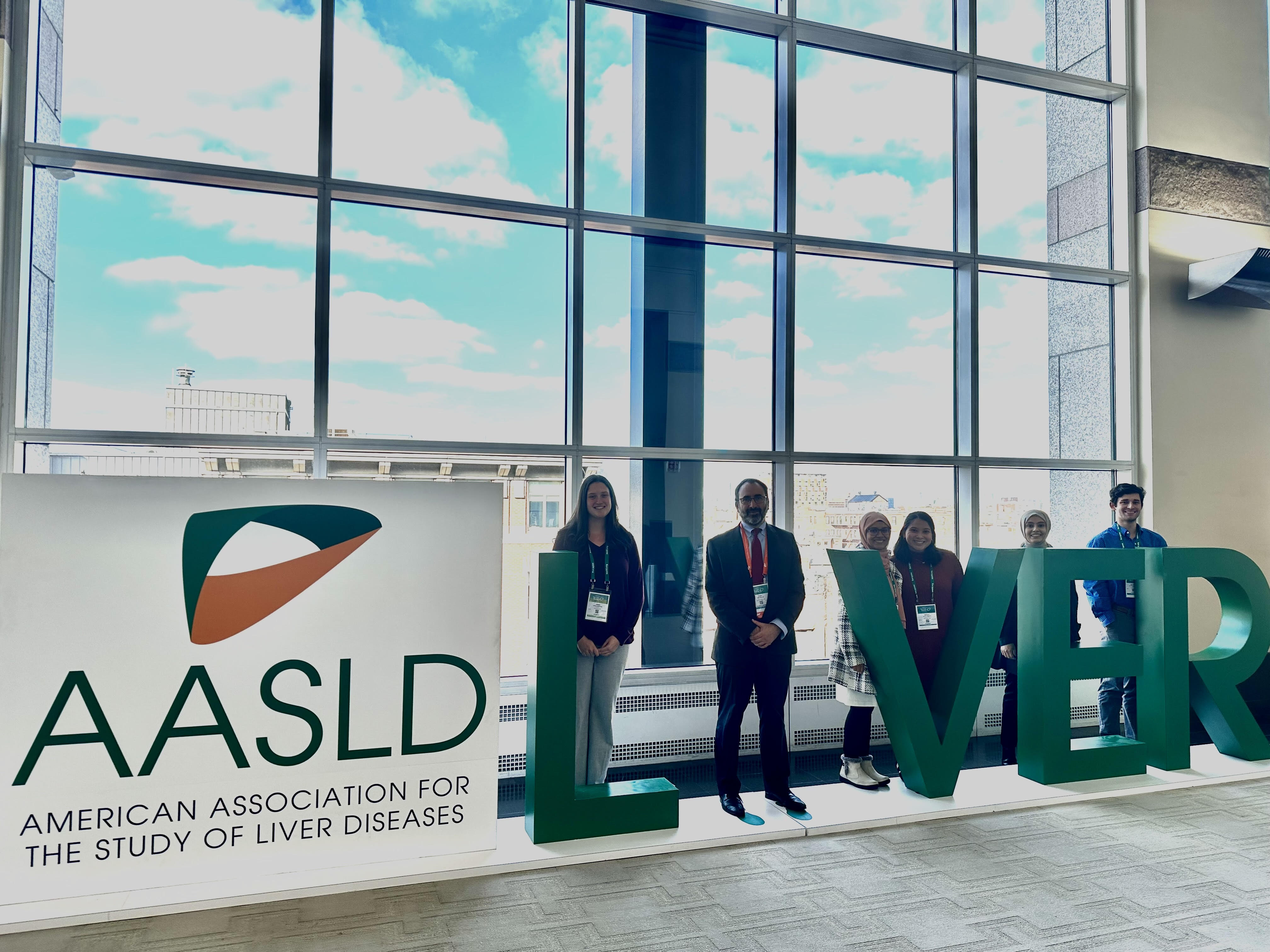 Lab Members stand in front of a sign at the American Association for the Study of Liver Diseases meeting.